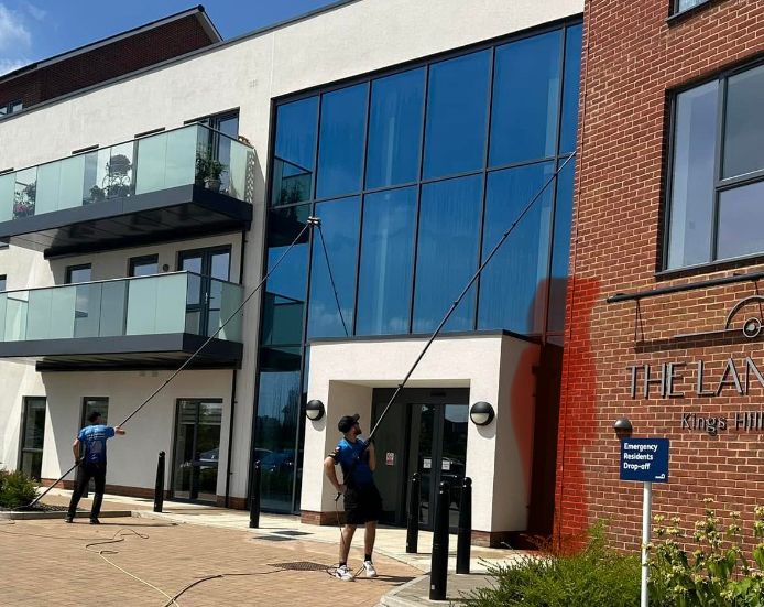 Two professionals performing commercial window cleaning on a modern building in Kent, featuring large glass windows and a brick façade, with water-fed poles for streak-free results.