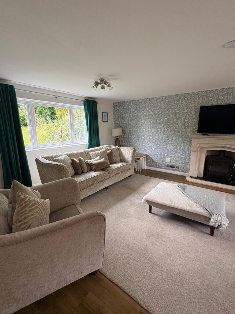 Living room interior with beige sofas, decorative cushions, and a coffee table, featuring a fireplace and large windows with green curtains, illustrating a clean and inviting space for end of tenancy cleaning services in Kent.